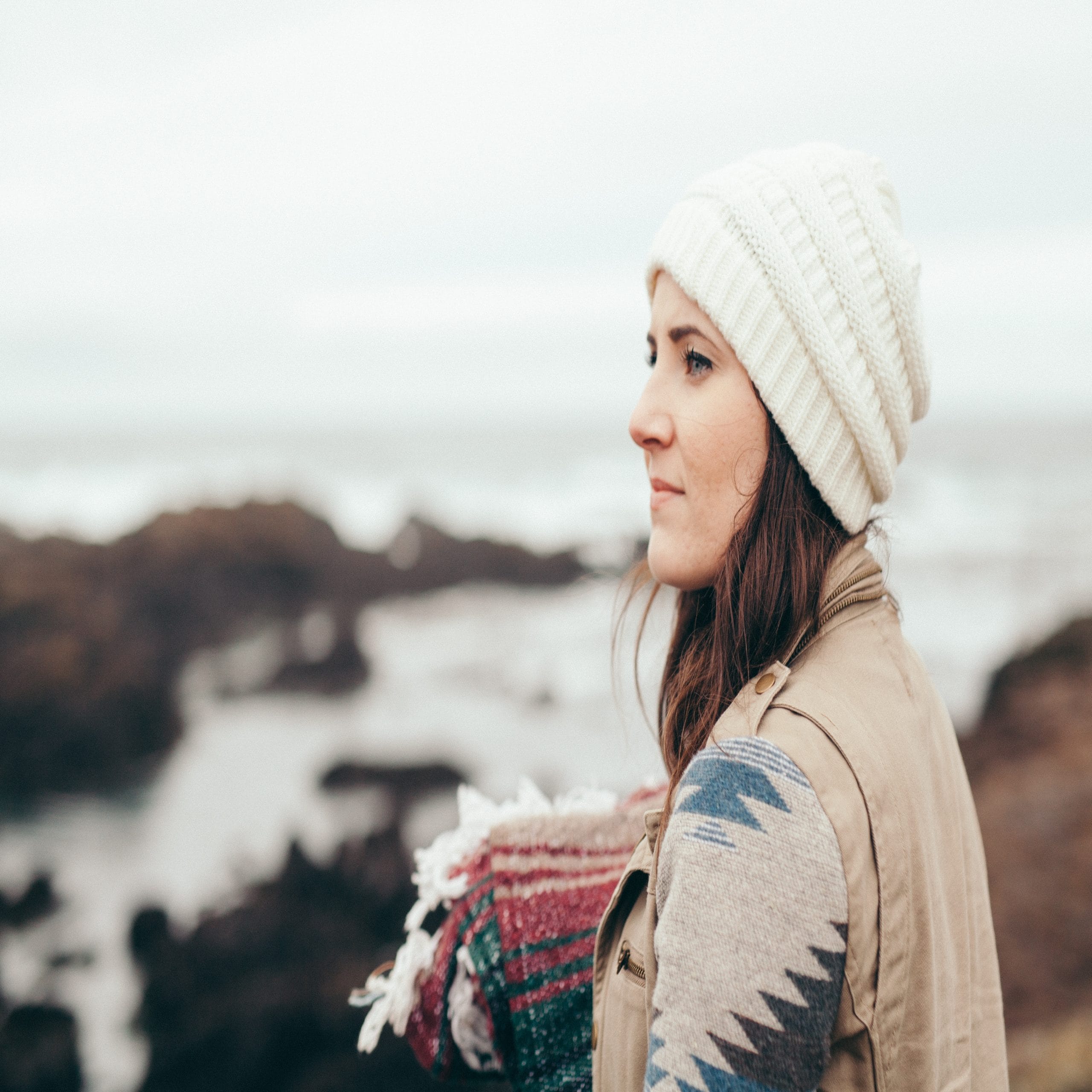 young-woman-looks-away-pondering-while-holding-a-blanket-by-the-ocean