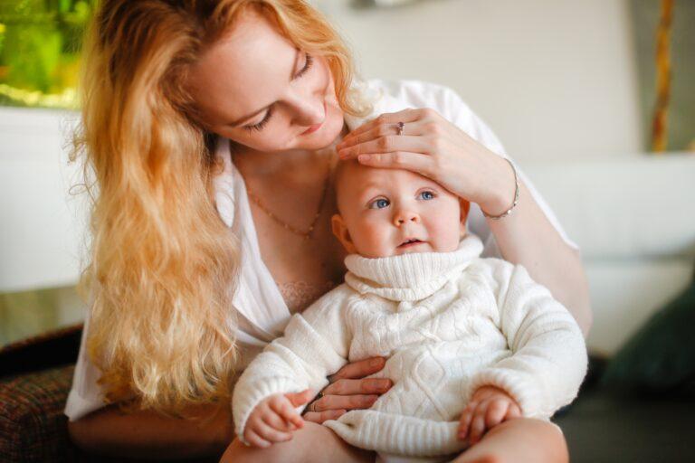 mother with long hair holds baby on her lap and places hand on baby's forehead - how to stay healthy when kids are sick