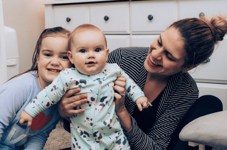 mom holding baby while toddler smiles beside her