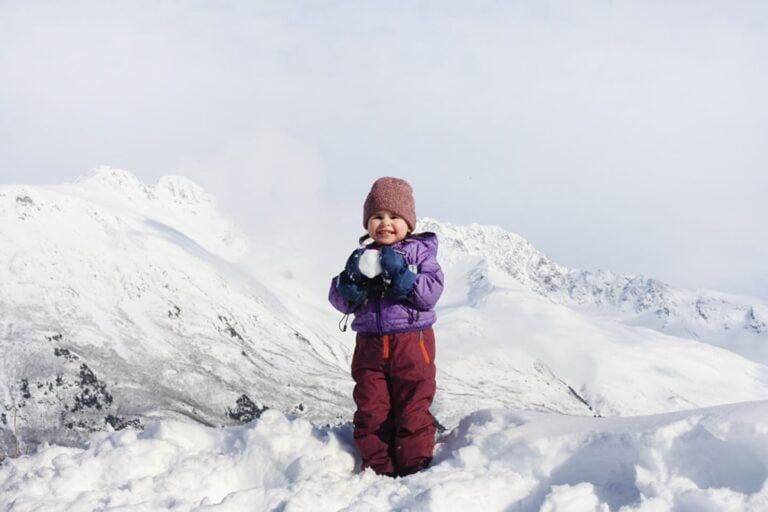 toddler holding a snow ball with mountains in the background - winter gear for kids