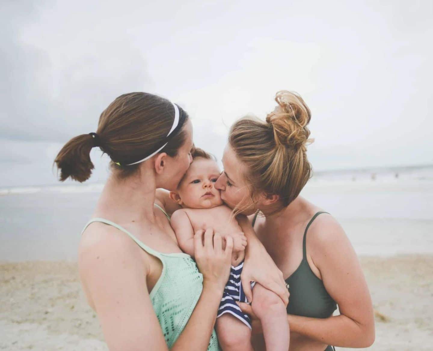 two women kissing a baby on the cheek on the beach