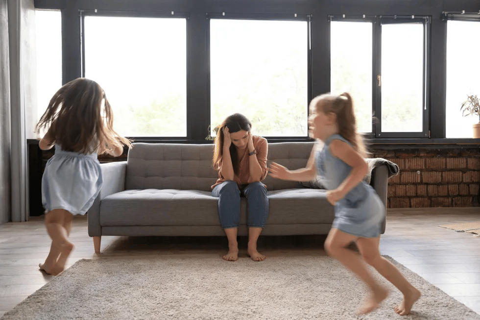 mother-sitting-on-sofa-with-children-running-around-house