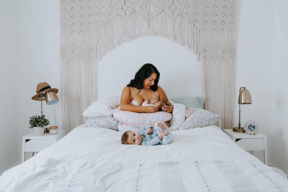 mom breastfeeding baby while another baby lays on the bed