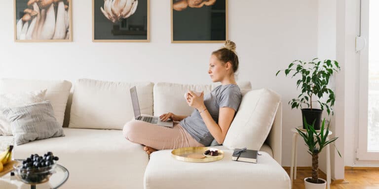 woman sitting on couch drinking coffee and thinking about toxic mom culture as she scrolls on her laptop