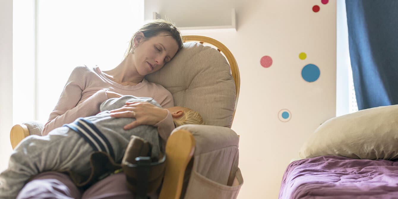 Exhausted mom sleeping with small child in a rocking chair.