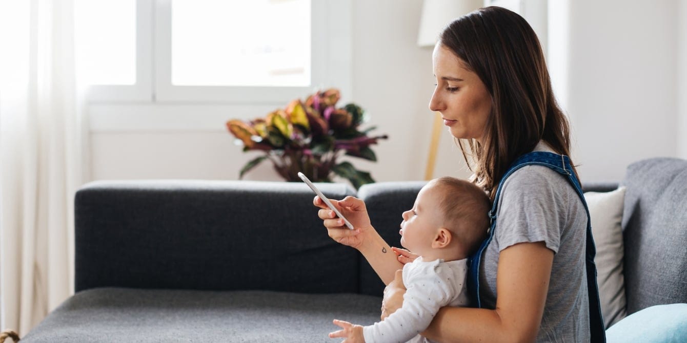 Young Mother Sitting With Her Baby Using Smartphone At Home - mom brain